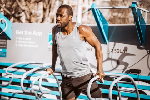 A man is performing a tricep dip exercise on parallel bars in an outdoor gym setting. He's wearing a gray tank top and black shorts. The background features workout equipment and a sign that suggests downloading an app.