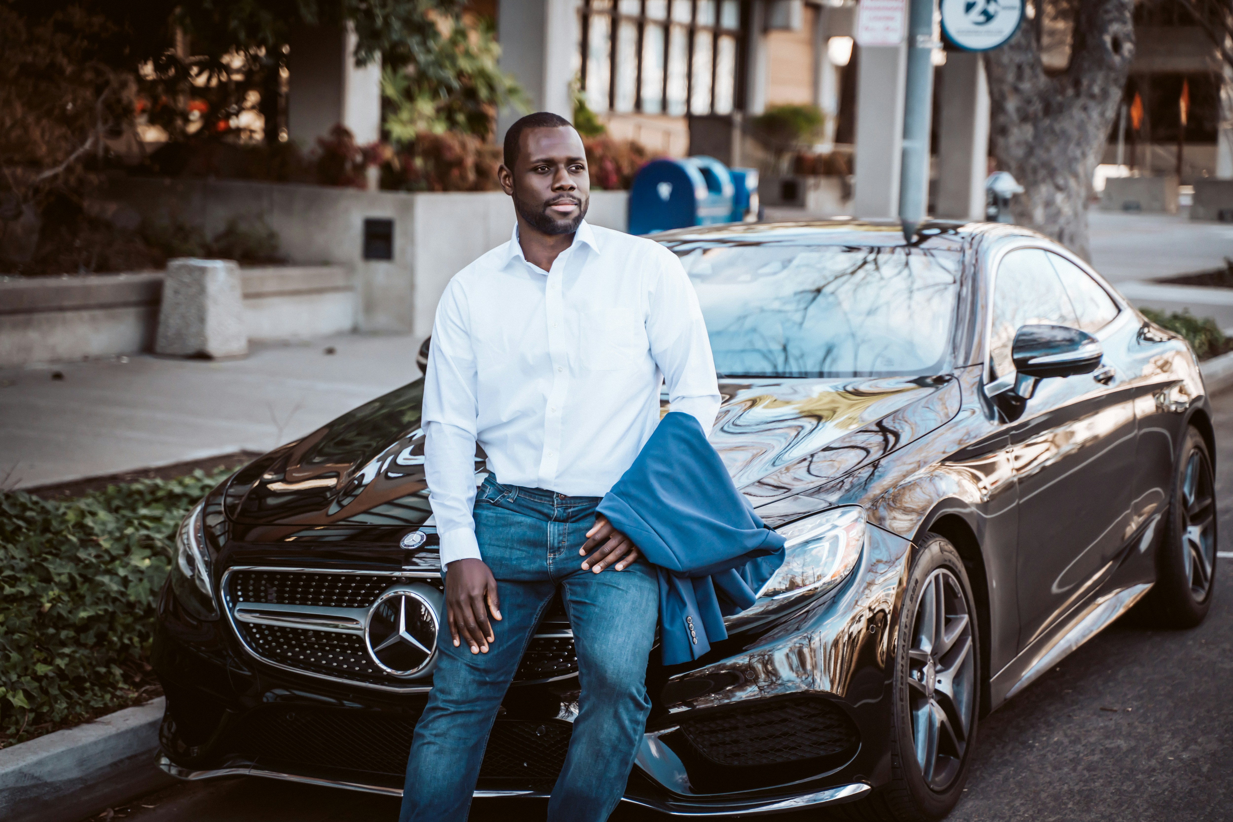 great photo recipe,how to photograph fortune vieyra dressed in business casual clothing and sitting on the hood of a mercedes with hand on jeans in the city..; man in white dress shirt and blue denim jeans standing beside black car during daytime
