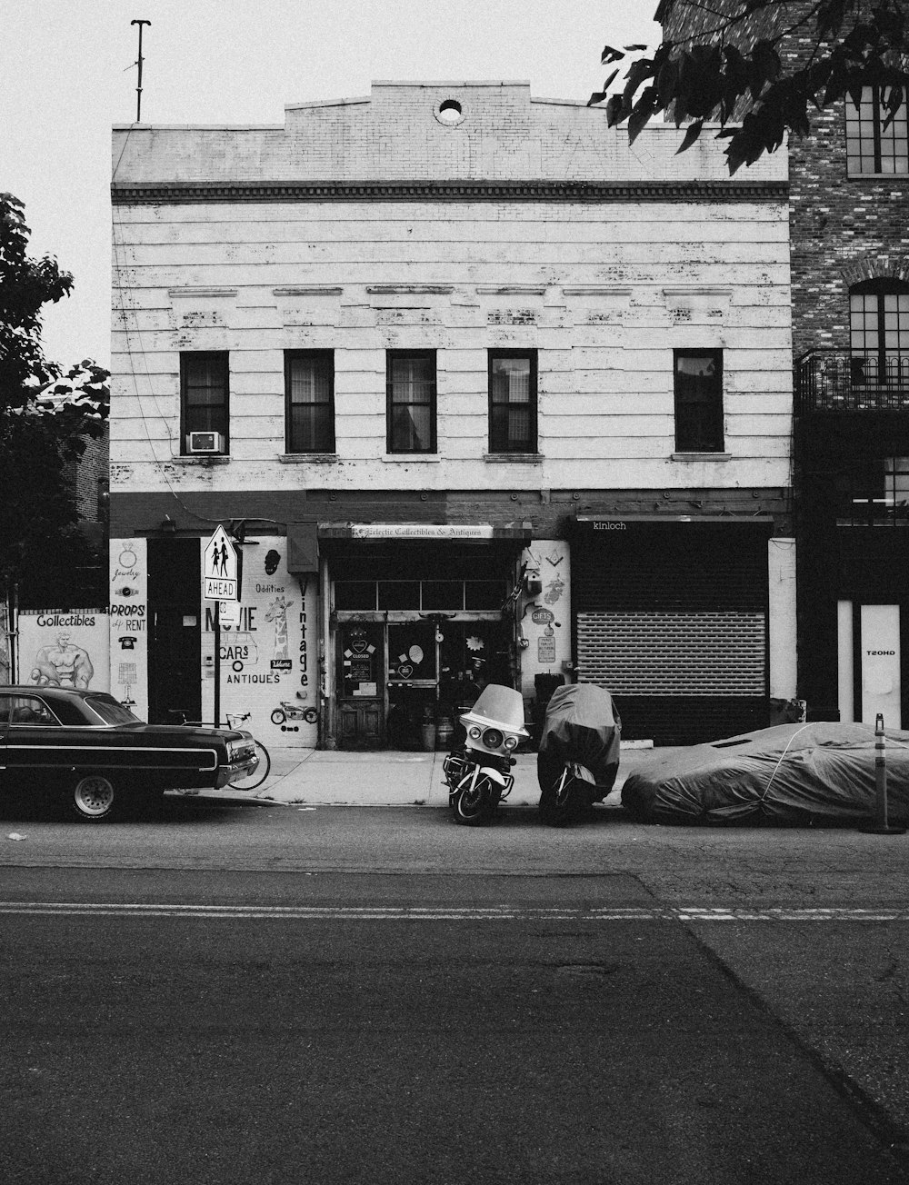 grayscale photo of man in black jacket and pants sitting on sidewalk near car parked beside