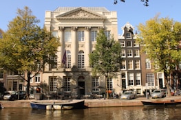 A grand neoclassical building with tall columns stands prominently by a canal. Surrounding the building are vibrant green trees and traditional Dutch canal houses with ornate facades. Several cars are parked along the canal, and small boats are moored by the water.