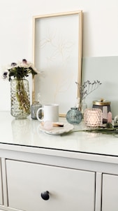 A minimalist tabletop scene featuring a white mug, a decorative vase with flowers, a framed line drawing, and various glass jars and candles. The arrangement includes a subtle mix of textures and pastel colors, with a focus on simplicity and elegance.