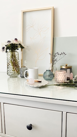 A minimalist tabletop scene featuring a white mug, a decorative vase with flowers, a framed line drawing, and various glass jars and candles. The arrangement includes a subtle mix of textures and pastel colors, with a focus on simplicity and elegance.