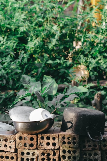 A rustic outdoor scene featuring a stack of bricks with various cooking pots and a white bowl placed on top. Lush green foliage in the background adds a natural contrast to the metallic kitchenware.