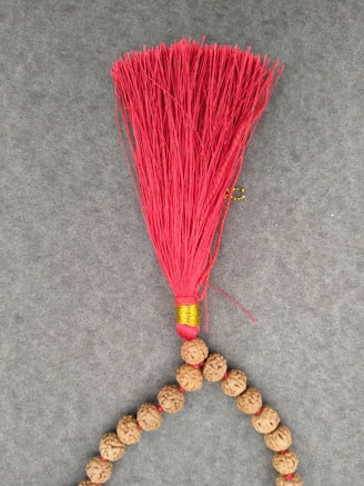 A string of rudraksha beads is arranged on a gray surface. At one end of the string, a vibrant red tassel is attached, wrapped with a golden thread at its base, adding a decorative element.