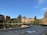 A scenic view of a university or college campus with historic buildings surrounding a large, circular reflecting pool. The architecture is characterized by brick structures with pointed roofs, and there are several trees and shrubs adding greenery. Some people are walking on the pathways, while others are sitting on benches near the pool. The sky is clear with a few clouds, and the setting is peaceful and serene.