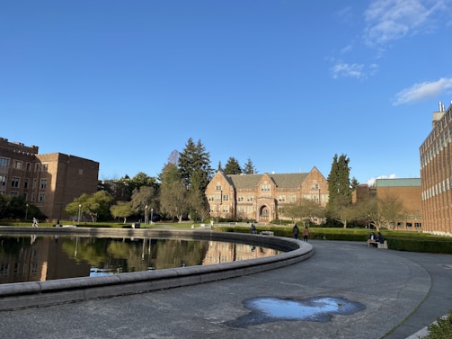 A scenic view of a university or college campus with historic buildings surrounding a large, circular reflecting pool. The architecture is characterized by brick structures with pointed roofs, and there are several trees and shrubs adding greenery. Some people are walking on the pathways, while others are sitting on benches near the pool. The sky is clear with a few clouds, and the setting is peaceful and serene.