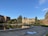 A scenic view of a university or college campus with historic buildings surrounding a large, circular reflecting pool. The architecture is characterized by brick structures with pointed roofs, and there are several trees and shrubs adding greenery. Some people are walking on the pathways, while others are sitting on benches near the pool. The sky is clear with a few clouds, and the setting is peaceful and serene.
