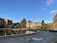 A scenic view of a university or college campus with historic buildings surrounding a large, circular reflecting pool. The architecture is characterized by brick structures with pointed roofs, and there are several trees and shrubs adding greenery. Some people are walking on the pathways, while others are sitting on benches near the pool. The sky is clear with a few clouds, and the setting is peaceful and serene.