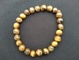 A bracelet made of round tiger eye beads arranged in a circular shape on a dark textured background. The beads exhibit a glossy finish with hues of brown, gold, and amber.