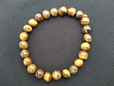 A bracelet made of round tiger eye beads arranged in a circular shape on a dark textured background. The beads exhibit a glossy finish with hues of brown, gold, and amber.