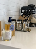 A kitchen counter featuring a candle in a glass jar, a bottle of liquid soap with a blue pump, three glass jars with metal lids in a black metal rack, a set of knives in a block, and a black stand mixer. The background shows a white tiled wall and an electrical outlet.