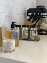 A kitchen counter featuring a candle in a glass jar, a bottle of liquid soap with a blue pump, three glass jars with metal lids in a black metal rack, a set of knives in a block, and a black stand mixer. The background shows a white tiled wall and an electrical outlet.