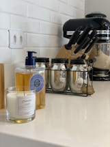 A kitchen counter featuring a candle in a glass jar, a bottle of liquid soap with a blue pump, three glass jars with metal lids in a black metal rack, a set of knives in a block, and a black stand mixer. The background shows a white tiled wall and an electrical outlet.
