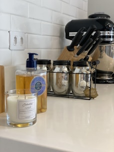 A kitchen counter featuring a candle in a glass jar, a bottle of liquid soap with a blue pump, three glass jars with metal lids in a black metal rack, a set of knives in a block, and a black stand mixer. The background shows a white tiled wall and an electrical outlet.