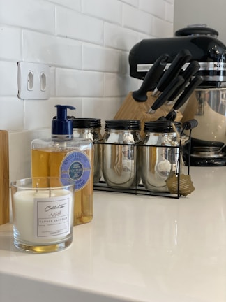 A kitchen counter featuring a candle in a glass jar, a bottle of liquid soap with a blue pump, three glass jars with metal lids in a black metal rack, a set of knives in a block, and a black stand mixer. The background shows a white tiled wall and an electrical outlet.