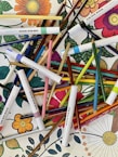 A vibrant assortment of colored pencils and markers arranged haphazardly over a coloring book page featuring detailed floral designs. Bright and varied colors create a lively and artistic atmosphere.