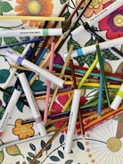 A vibrant assortment of colored pencils and markers arranged haphazardly over a coloring book page featuring detailed floral designs. Bright and varied colors create a lively and artistic atmosphere.