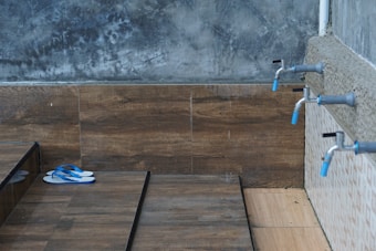 A tiled wash area with three water taps lined along a concrete wall. A pair of blue flip flops is placed on the wooden floor surface with a textured gray wall in the background.