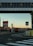 An urban setting featuring a transportation hub with a bridge overhead. Several buses are visible, and signs indicate lanes for authorized vehicles and buses only. The area is marked by traffic lines and street signs, and the sky has a warm, sunset glow.