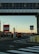 An urban setting featuring a transportation hub with a bridge overhead. Several buses are visible, and signs indicate lanes for authorized vehicles and buses only. The area is marked by traffic lines and street signs, and the sky has a warm, sunset glow.