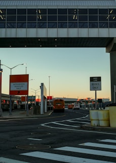 An urban setting featuring a transportation hub with a bridge overhead. Several buses are visible, and signs indicate lanes for authorized vehicles and buses only. The area is marked by traffic lines and street signs, and the sky has a warm, sunset glow.