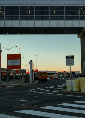 An urban setting featuring a transportation hub with a bridge overhead. Several buses are visible, and signs indicate lanes for authorized vehicles and buses only. The area is marked by traffic lines and street signs, and the sky has a warm, sunset glow.