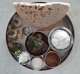 A traditional Indian meal served on a round metal plate featuring a variety of dishes. It includes rice, a curry with a dark sauce garnished with herbs, a large piece of flatbread, sliced vegetables or rolls with herbs, a small bowl of chopped green salad, a bowl of white salad, and two smaller bowls with liquid dishes. Lemon wedges and a spoon are also present.