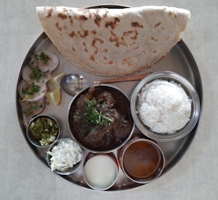 A traditional Indian meal served on a round metal plate featuring a variety of dishes. It includes rice, a curry with a dark sauce garnished with herbs, a large piece of flatbread, sliced vegetables or rolls with herbs, a small bowl of chopped green salad, a bowl of white salad, and two smaller bowls with liquid dishes. Lemon wedges and a spoon are also present.