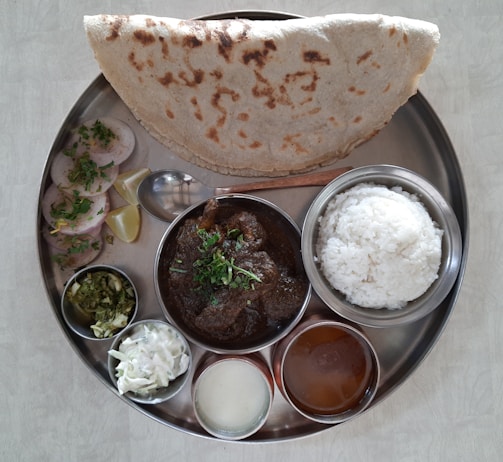 A traditional Indian meal served on a round metal plate featuring a variety of dishes. It includes rice, a curry with a dark sauce garnished with herbs, a large piece of flatbread, sliced vegetables or rolls with herbs, a small bowl of chopped green salad, a bowl of white salad, and two smaller bowls with liquid dishes. Lemon wedges and a spoon are also present.