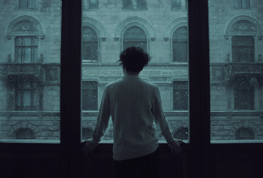 A young woman standing quietly at a window, surrounded by people in her life and yet still without anyone to think out loud with