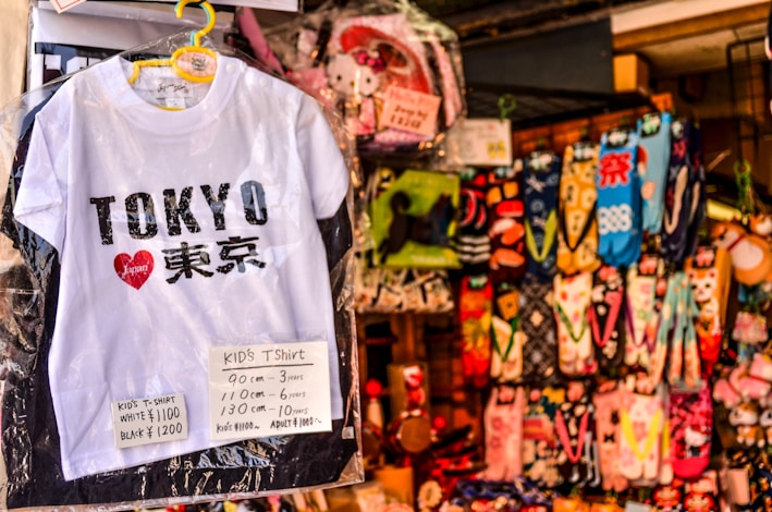 A white children's T-shirt with 'Tokyo' written on it in bold black letters, accompanied by a red heart logo containing the word 'Japan'. Price tags in yen specify sizes and costs. Behind the T-shirt, there's a colorful array of hanging socks featuring various patterns and designs.