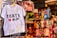 A white children's T-shirt with 'Tokyo' written on it in bold black letters, accompanied by a red heart logo containing the word 'Japan'. Price tags in yen specify sizes and costs. Behind the T-shirt, there's a colorful array of hanging socks featuring various patterns and designs.