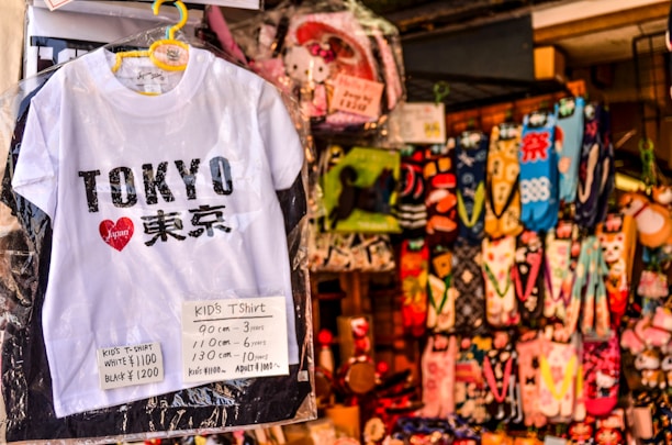A white children's T-shirt with 'Tokyo' written on it in bold black letters, accompanied by a red heart logo containing the word 'Japan'. Price tags in yen specify sizes and costs. Behind the T-shirt, there's a colorful array of hanging socks featuring various patterns and designs.