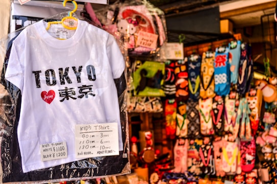A white children's T-shirt with 'Tokyo' written on it in bold black letters, accompanied by a red heart logo containing the word 'Japan'. Price tags in yen specify sizes and costs. Behind the T-shirt, there's a colorful array of hanging socks featuring various patterns and designs.