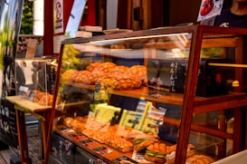 A bakery display case filled with a variety of baked goods, including bread and pastries. The baked items are arranged neatly on wooden shelves behind a glass pane. Signage written in Japanese is visible, indicating the products and pricing. The setting appears to be a street-side bakery with a warm and inviting atmosphere.