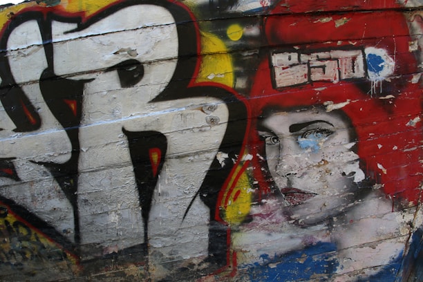 Graffiti art covering an urban wall, featuring bold, abstract lettering and a striking face of a woman with red hair and a serious expression. The artwork includes bright colors and a textured surface with peeling paint.