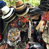 A vibrant display of assorted items, including neatly stacked hats of various materials and colors, colorful alphabet beads, decorative hair accessories, jewelry, and fabric belts. The collection appears densely packed and visually stimulating, likely part of a market stall.
