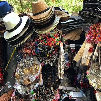 A vibrant display of assorted items, including neatly stacked hats of various materials and colors, colorful alphabet beads, decorative hair accessories, jewelry, and fabric belts. The collection appears densely packed and visually stimulating, likely part of a market stall.