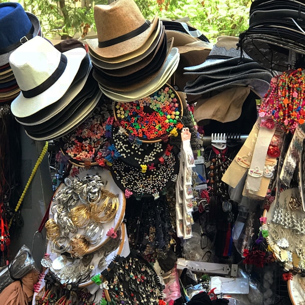 A vibrant display of assorted items, including neatly stacked hats of various materials and colors, colorful alphabet beads, decorative hair accessories, jewelry, and fabric belts. The collection appears densely packed and visually stimulating, likely part of a market stall.