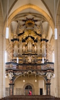 An ornate pipe organ is mounted against the wall of a large cathedral interior, featuring intricate carvings, golden accents, and towering pipes. Below the organ, a single figure in a red coat walks along wooden pews. The architectural design includes grand arches and detailed sculptures.