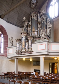 A beautifully ornate pipe organ is positioned prominently in a church interior. The organ features intricate carvings and is adorned with statues. Below the organ, there are rows of wooden pews and a simple altar. The walls are light-colored, and there are large arched windows allowing natural light to flood the space.