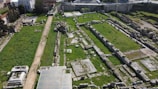 The image depicts an archaeological site with ruins, featuring remnants of stone walls and columns. The area is covered with patches of green grass and is bordered by a dirt path. Trees and modern buildings are visible in the background.