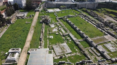 The image depicts an archaeological site with ruins, featuring remnants of stone walls and columns. The area is covered with patches of green grass and is bordered by a dirt path. Trees and modern buildings are visible in the background.