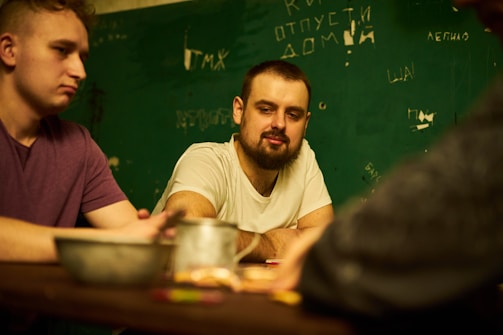 Three people are seated around a table in a dimly lit room, engaged in conversation. The back wall has scribbled writings in Cyrillic script, suggesting a casual or informal setting. The individuals appear relaxed, and there are cups and possibly snacks on the table.