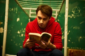 A young man is sitting on a metal bunk bed in a room with green walls covered in graffiti. He is wearing a red hoodie and dark pants, and is engrossed in reading a book.