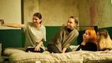 Four young women are sitting together on a bed inside what appears to be a room with textured walls. One woman on the left is taking a selfie with a smartphone, while the others are looking towards the camera, seemingly enjoying the moment.