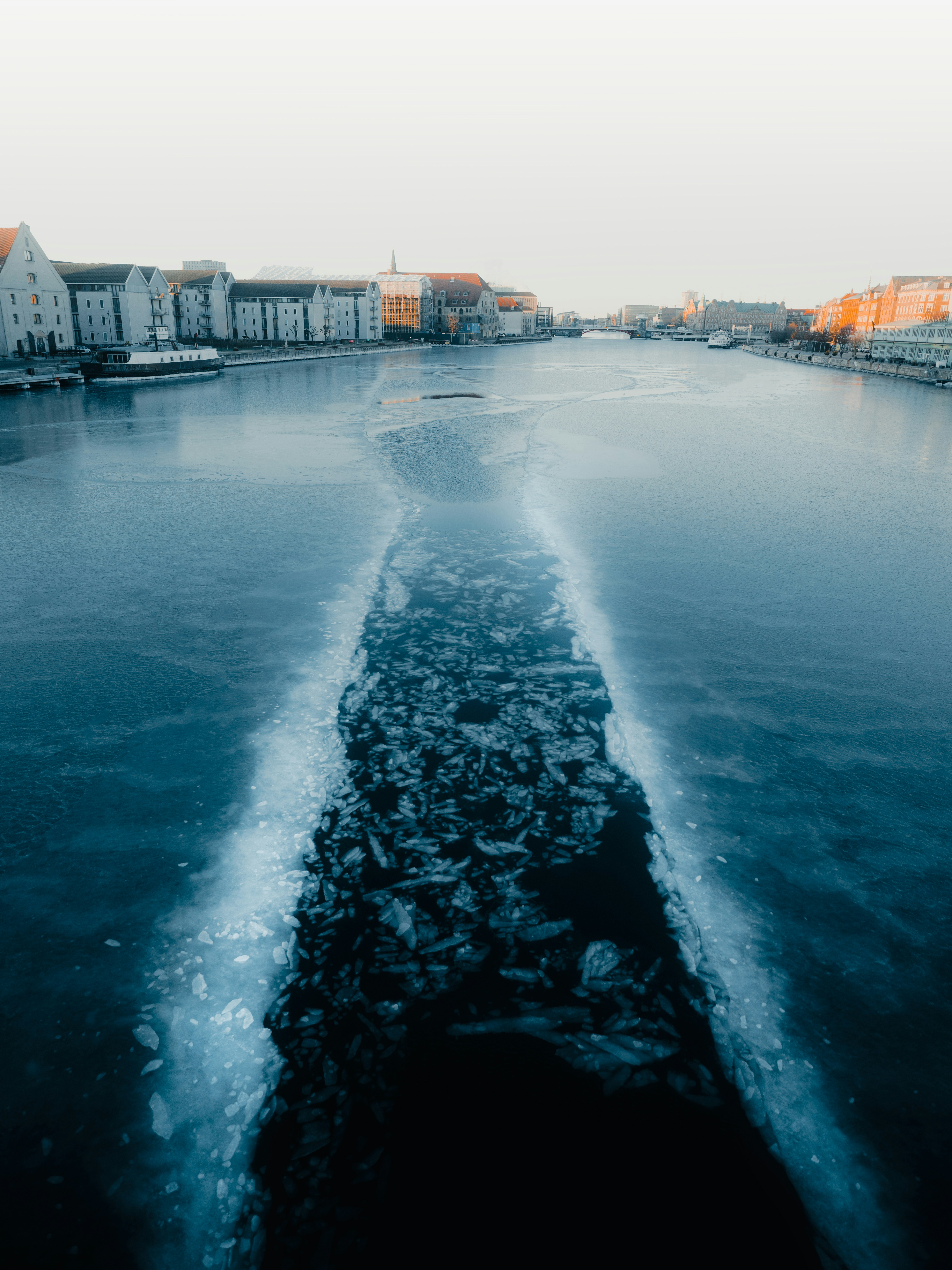 A channel cut through ice on a wide harbour, low warehouses and pale facades along both banks