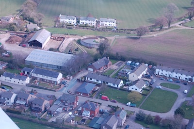 An aerial view of a rural area featuring residential buildings with pitched roofs arranged in orderly rows and clusters. A large barn and several agricultural structures are visible, along with open fields and grassy areas. The scene is surrounded by trees and shrubs, suggesting a countryside setting with a mix of modern and traditional architecture.