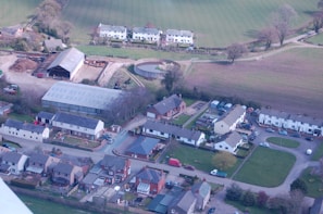 An aerial view of a rural area featuring residential buildings with pitched roofs arranged in orderly rows and clusters. A large barn and several agricultural structures are visible, along with open fields and grassy areas. The scene is surrounded by trees and shrubs, suggesting a countryside setting with a mix of modern and traditional architecture.