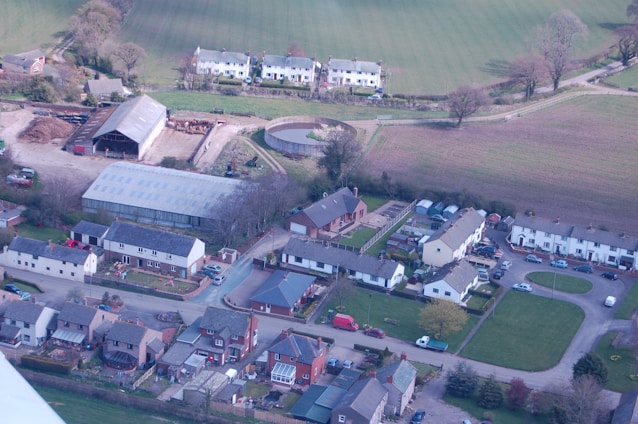 An aerial view of a rural area featuring residential buildings with pitched roofs arranged in orderly rows and clusters. A large barn and several agricultural structures are visible, along with open fields and grassy areas. The scene is surrounded by trees and shrubs, suggesting a countryside setting with a mix of modern and traditional architecture.
